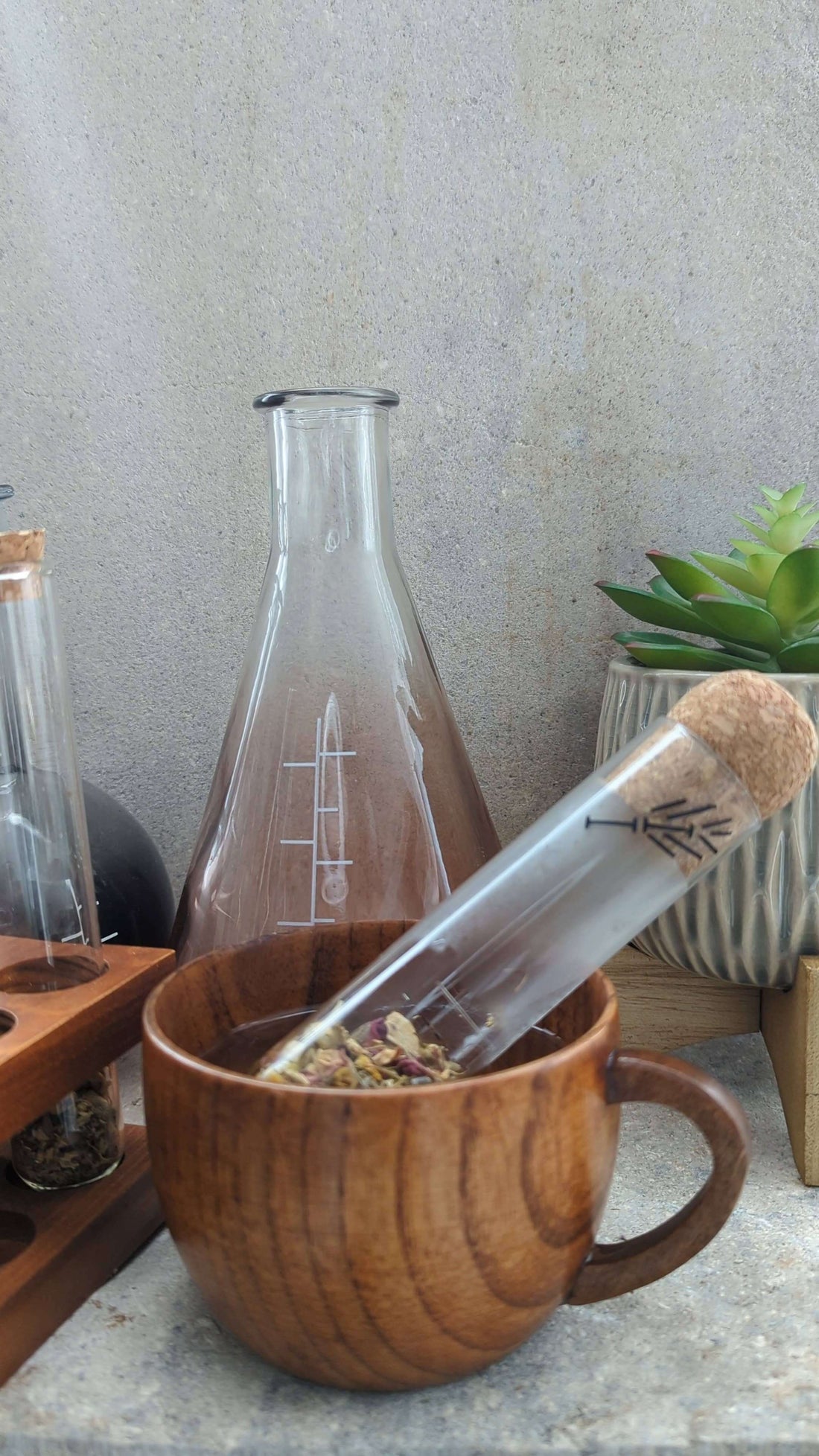 Glass test tube tea infuser resting in a wooden cup, surrounded by elegant glassware and a plant.